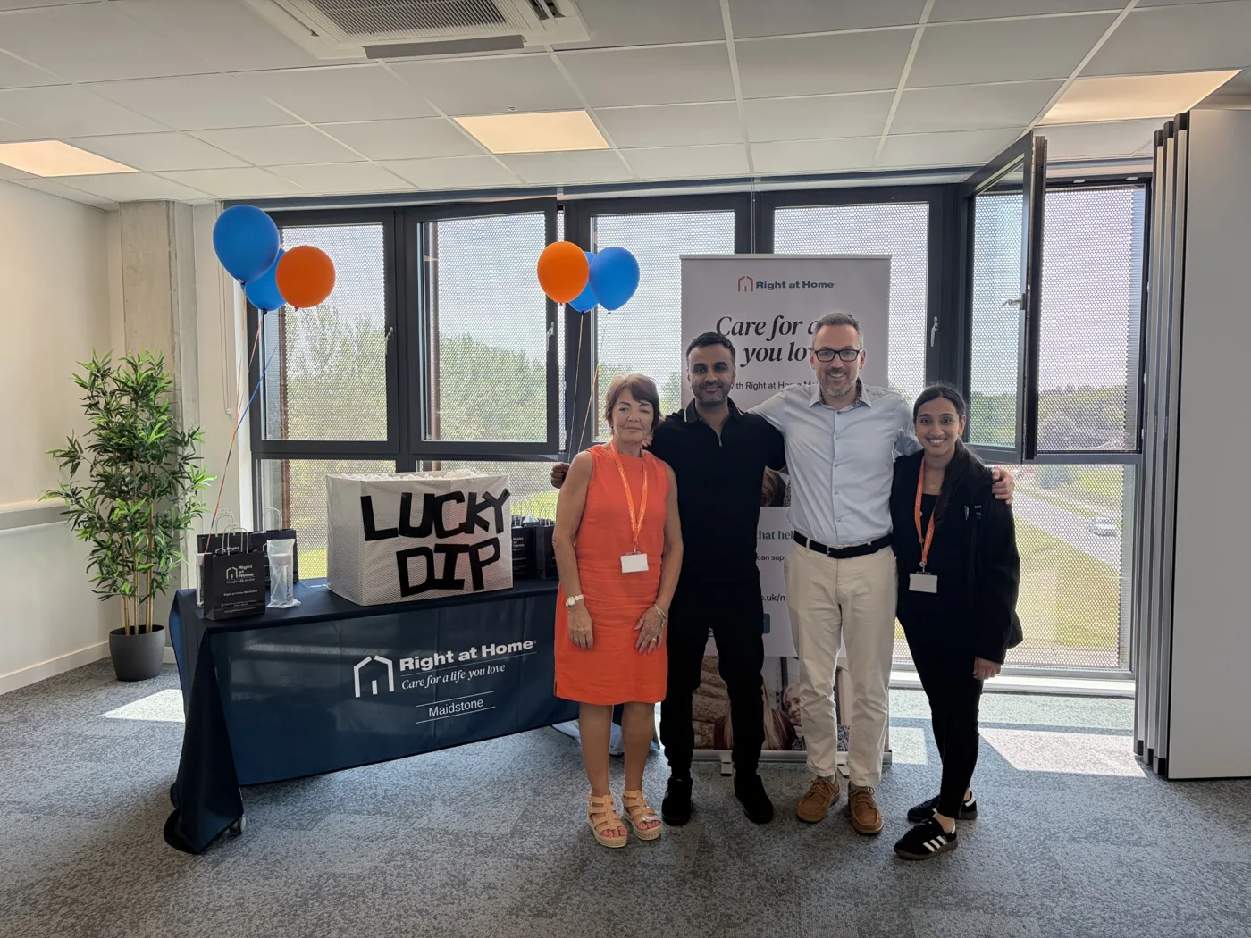 Four people standing together in a bright office in front of a Right at Home banner, beside a table with a Lucky Dip box, gift bags and blue and orange balloons